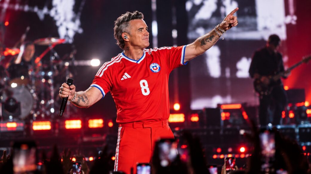 Un show de estadio nocturno tipo concierto de pop/rock británico, con el artista en el centro del escenario bajo luces de espectáculo y una multitud visible, evocando el recital de Robbie Williams en el Bicentenario de La Florida sin reclamar ser una foto real del evento.