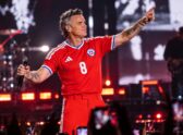 Un show de estadio nocturno tipo concierto de pop/rock británico, con el artista en el centro del escenario bajo luces de espectáculo y una multitud visible, evocando el recital de Robbie Williams en el Bicentenario de La Florida sin reclamar ser una foto real del evento.
