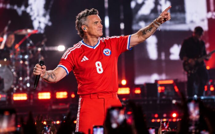 Un show de estadio nocturno tipo concierto de pop/rock británico, con el artista en el centro del escenario bajo luces de espectáculo y una multitud visible, evocando el recital de Robbie Williams en el Bicentenario de La Florida sin reclamar ser una foto real del evento.